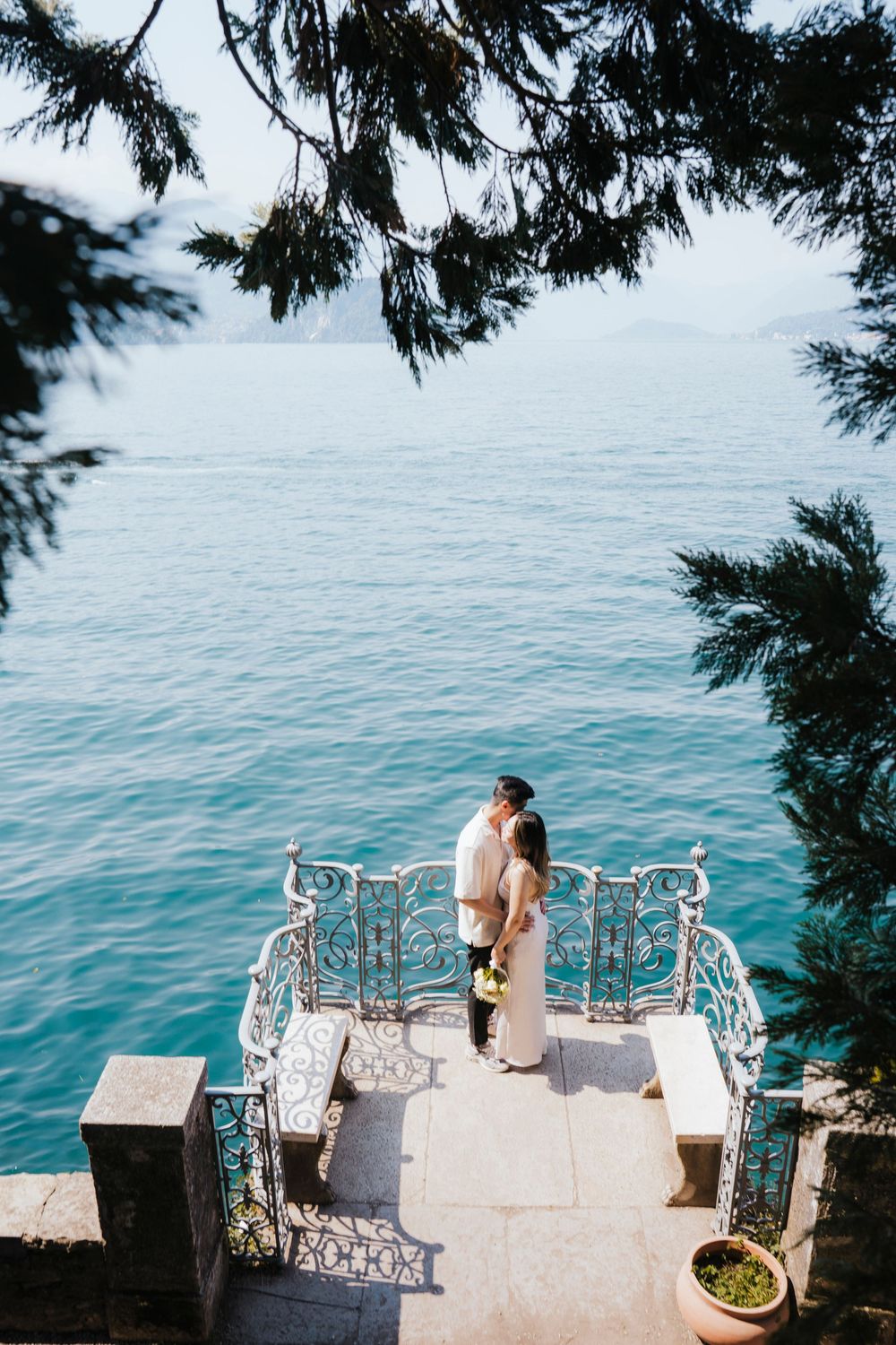 A romantic couple sits on a stone balcony overlooking the turquoise Mediterranean Sea through ornate white railings.