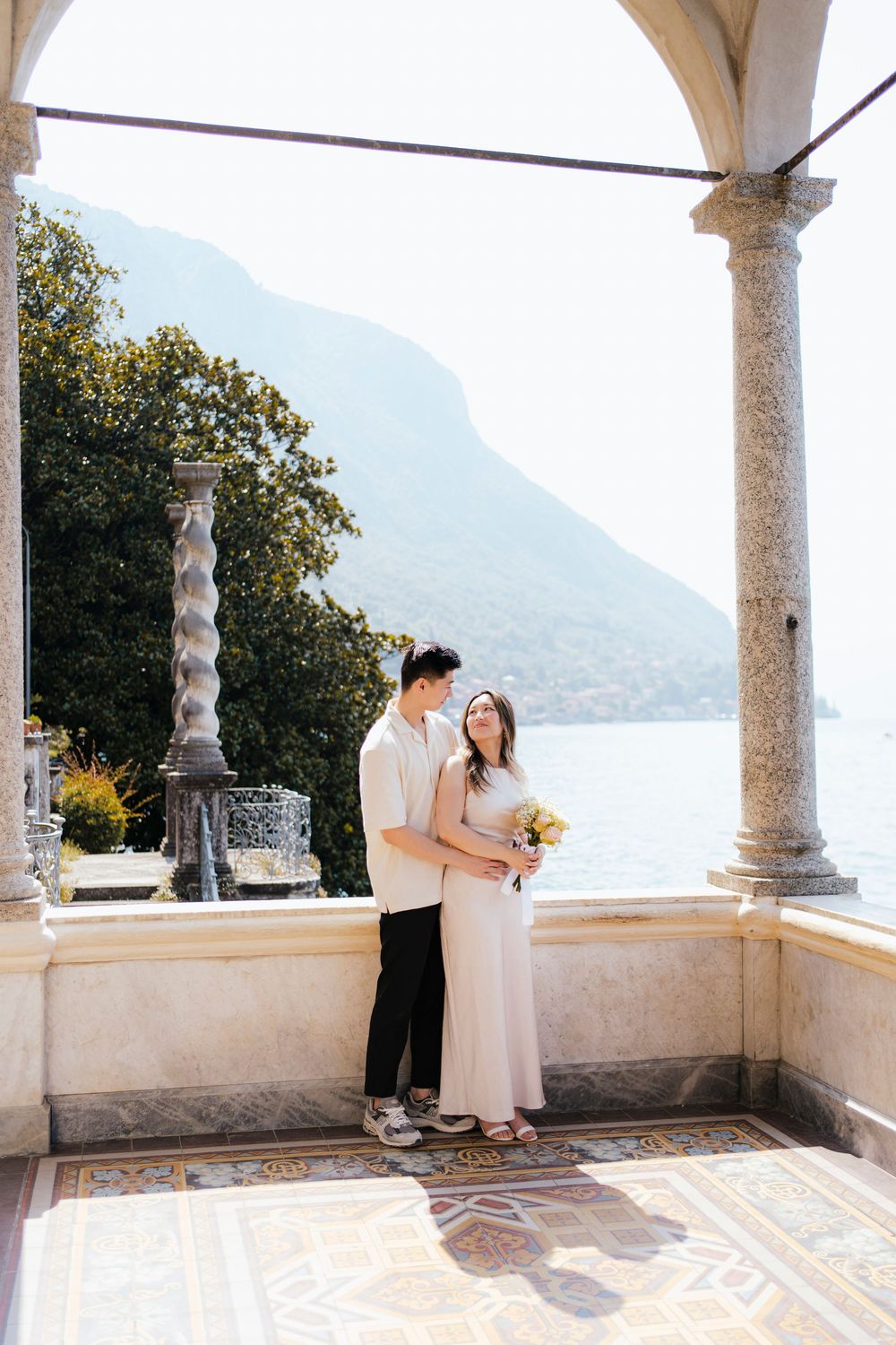 A couple shares an intimate moment on a marble terrace overlooking a scenic lake and mountains in Italy.