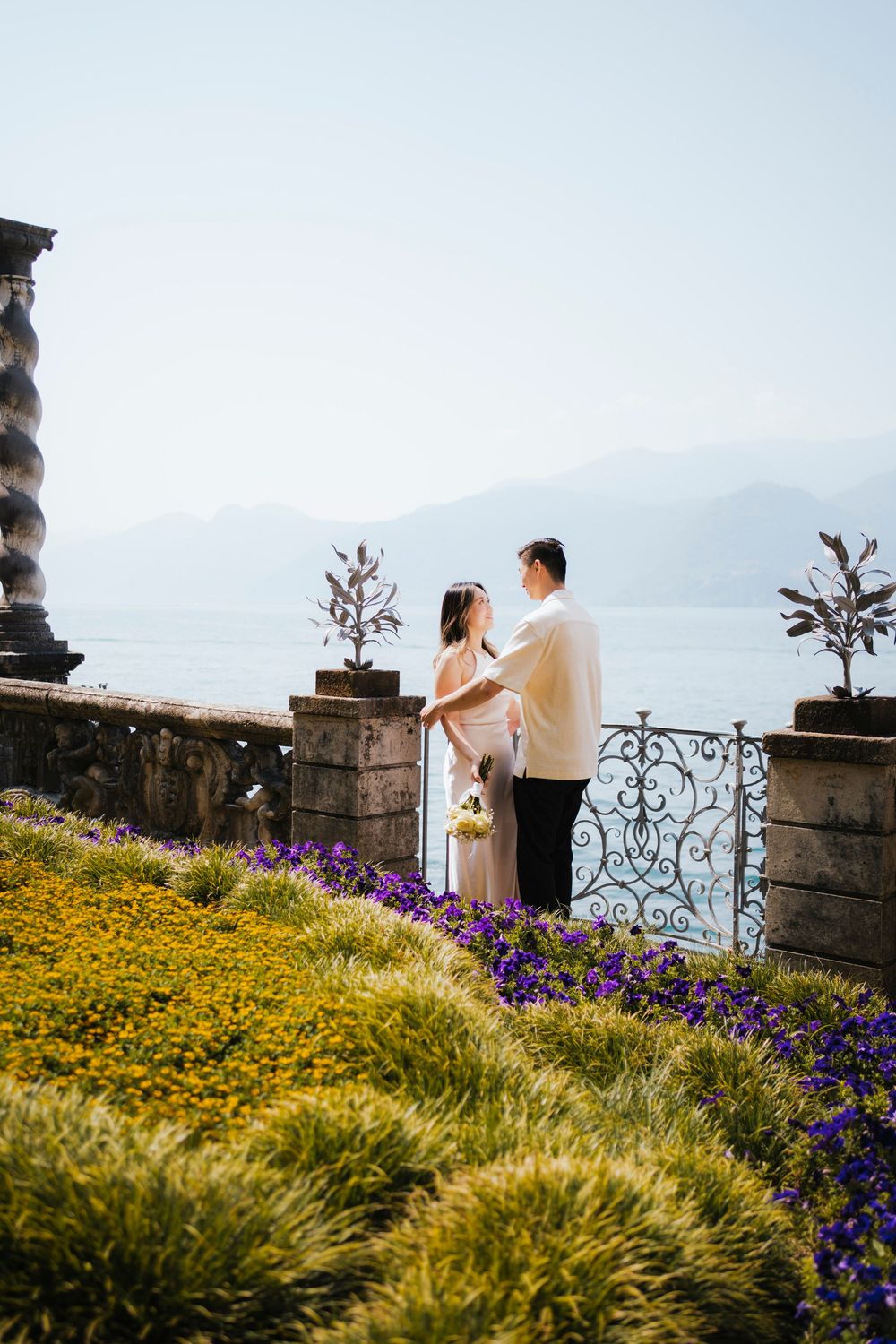 A romantic moment captured in a garden terrace with yellow and purple flowers overlooking a misty lake and mountains.