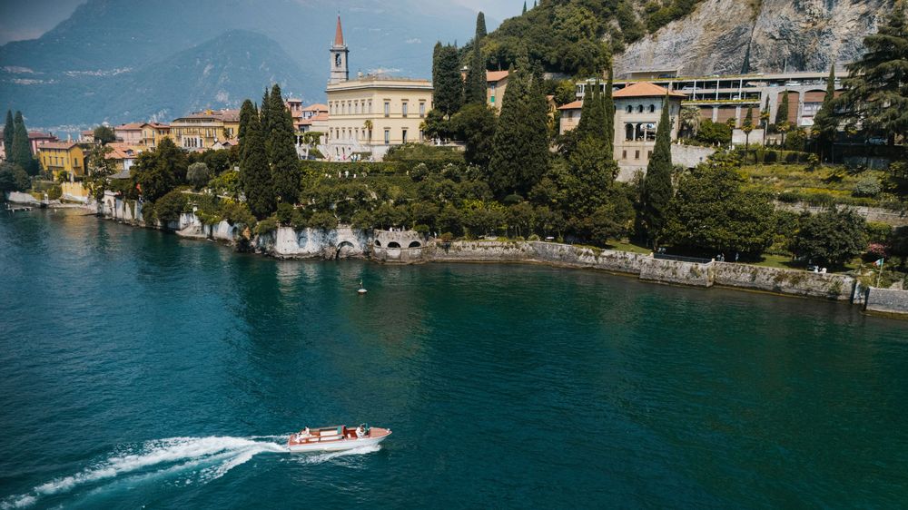 Speedboat cruising past lakeside buildings and cypress trees on Lake Como with church tower in background.