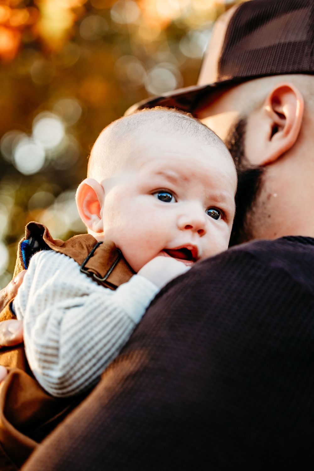 A baby in a gray sweater looks over someone's shoulder with warm autumn bokeh lighting in the background.