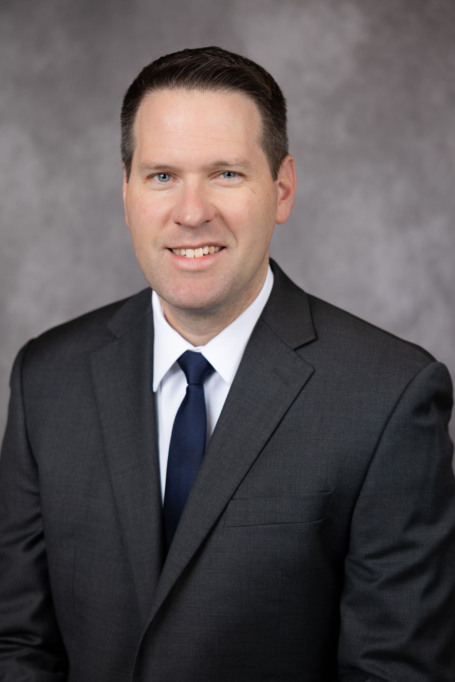 Professional headshot portrait of person in dark suit and navy tie against a gray studio background.