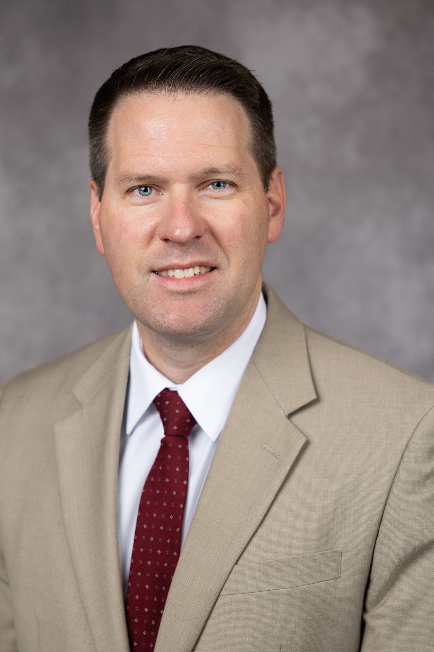 Professional headshot of a person wearing a tan suit jacket and red polka dot tie against a gray background.