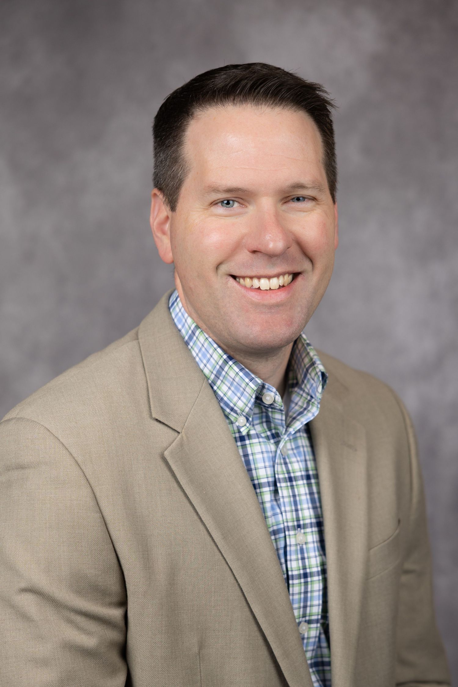 Professional headshot portrait of person in beige blazer and blue checkered button-down shirt against gray background.