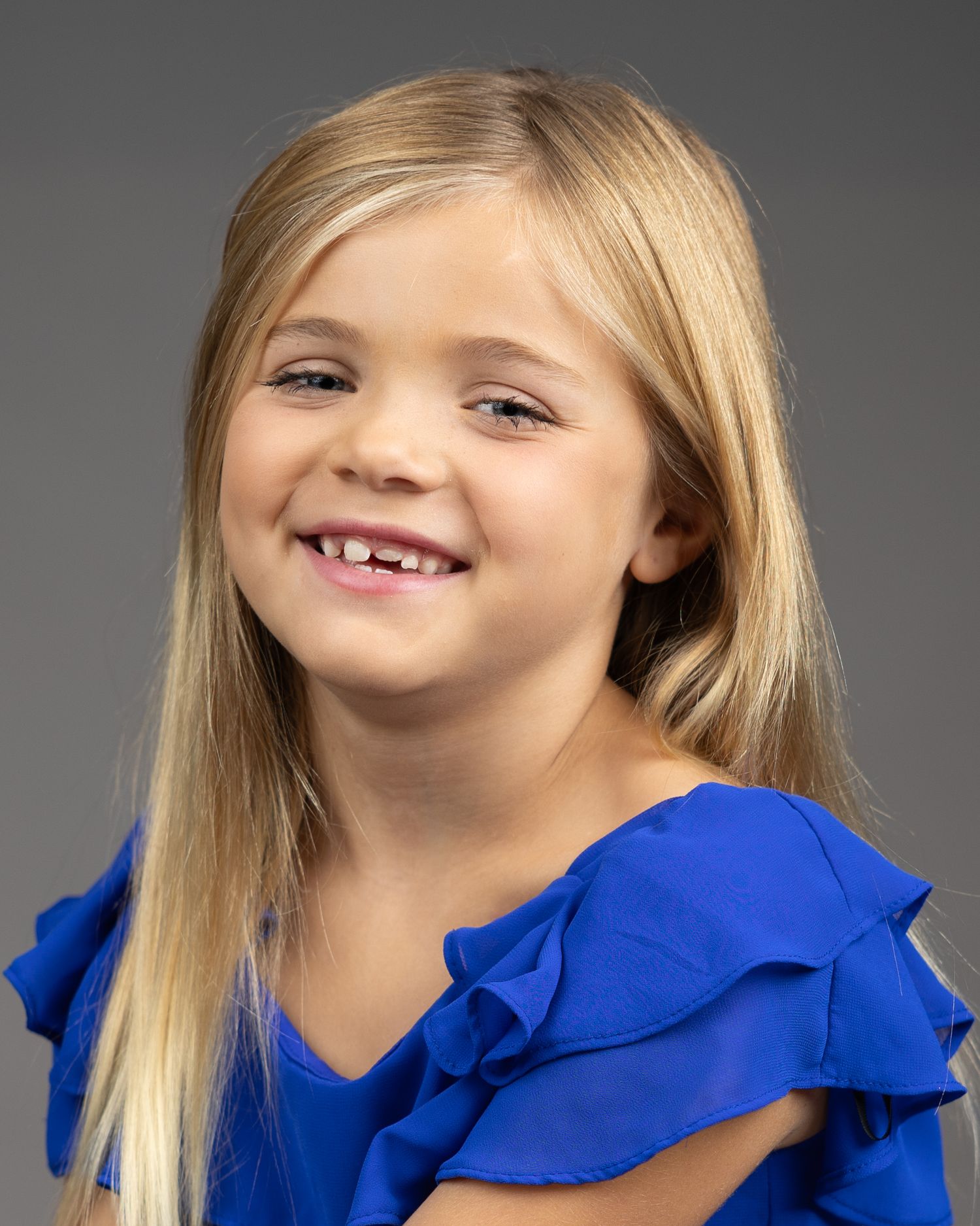 Young student in blue ruffled sleeve top poses for school portrait against grey background.