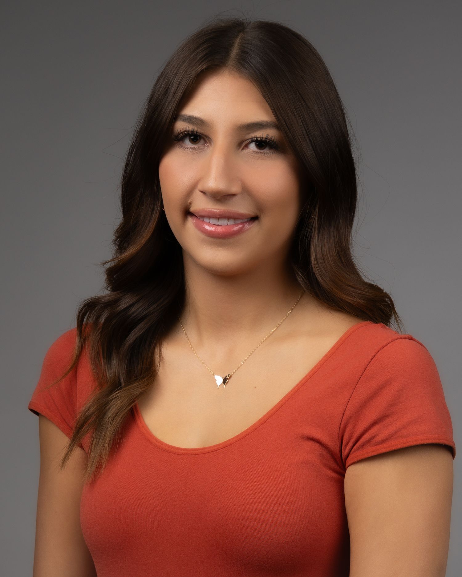 Professional headshot of woman in orange top with wavy brown hair against grey backdrop.