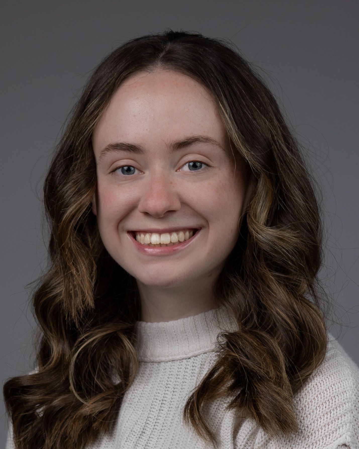 Headshot of woman in white sweater with wavy brown hair and bright smile.