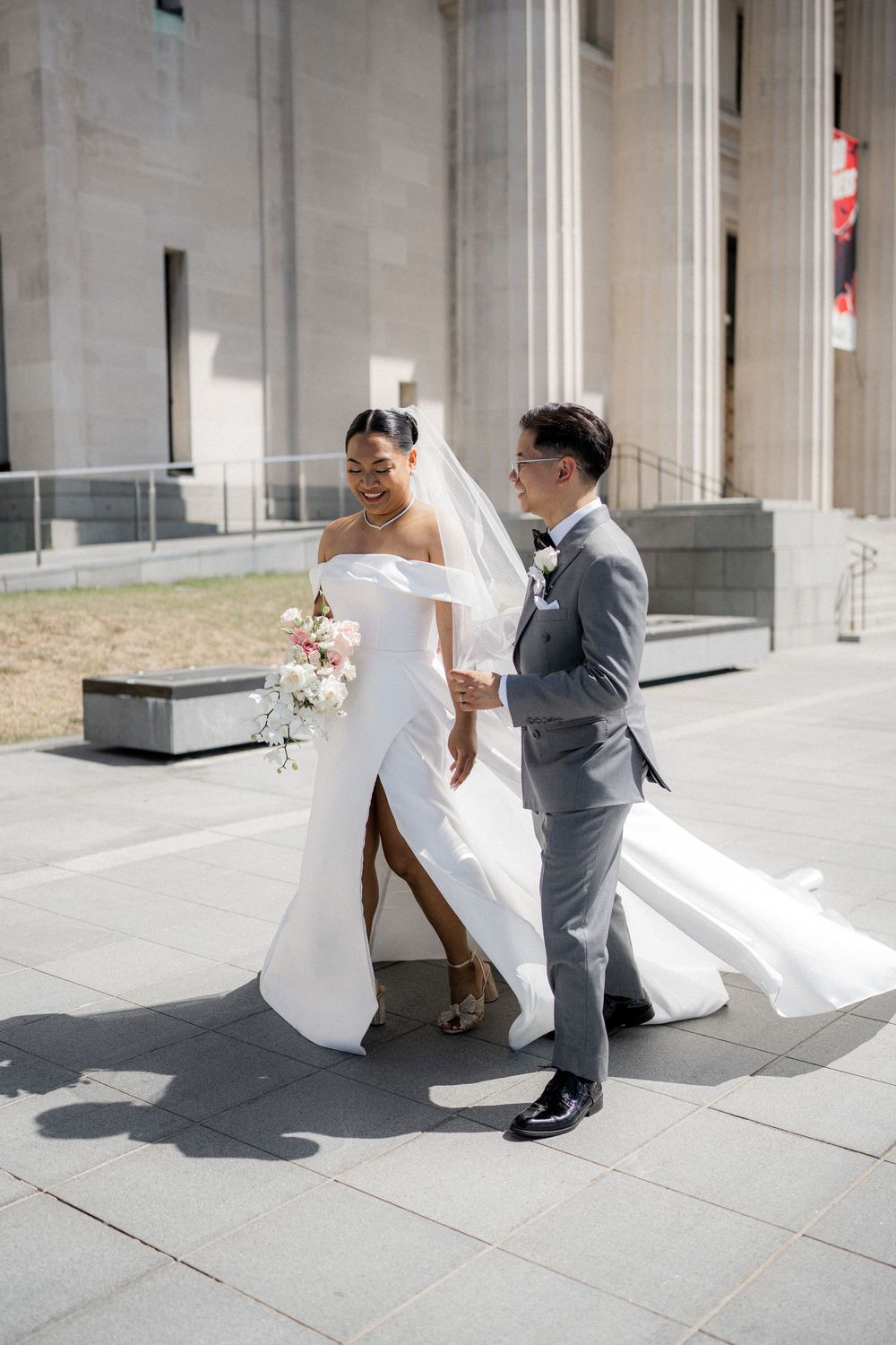 A couple walks along a stone building exterior during their wedding day.
