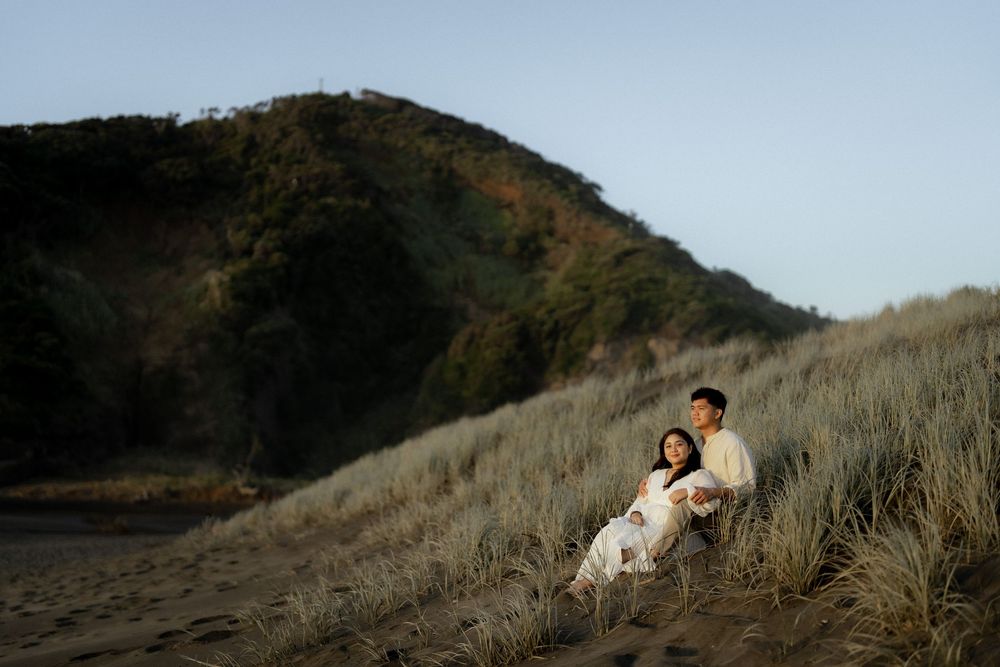 A couple sits together on a grassy hillside during a romantic outdoor photoshoot at sunset.