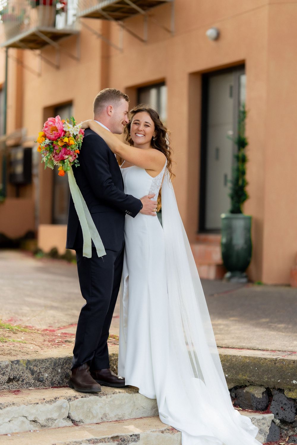 Wedding couple poses for romantic photos against a peach-colored building wall.