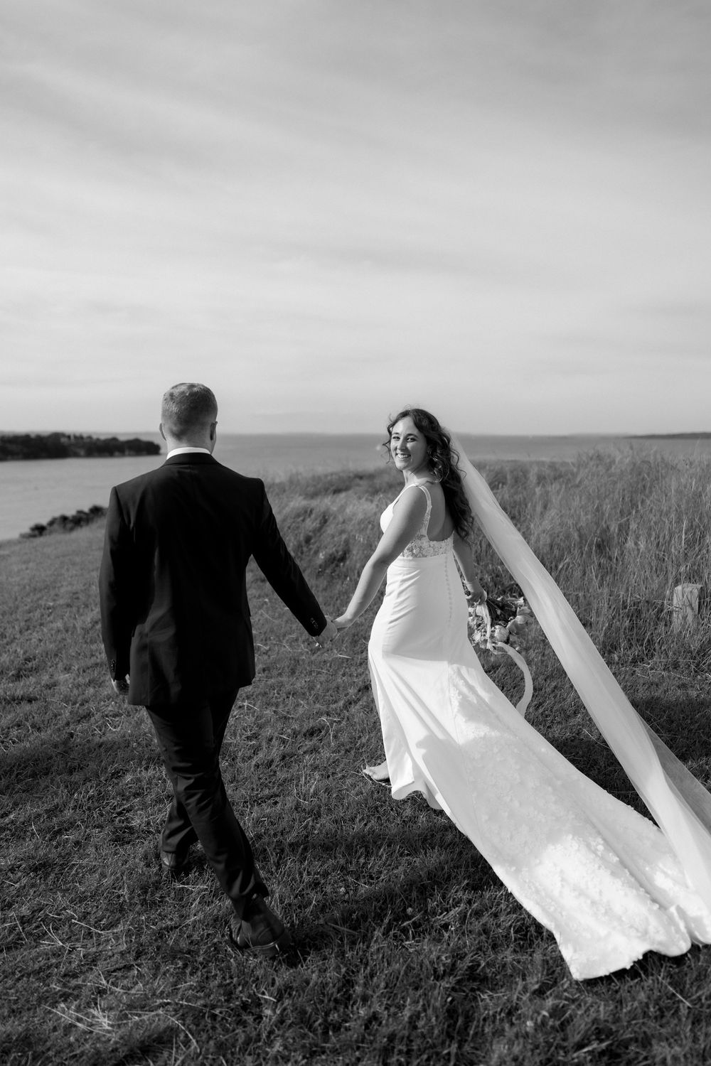 Black and white photo of newlyweds walking dramatically across a windswept coastal field.