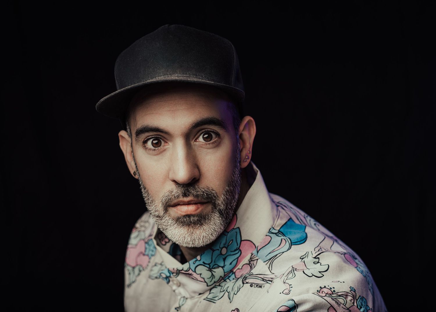 Studio portrait with patterned shirt and black hat against dark background.