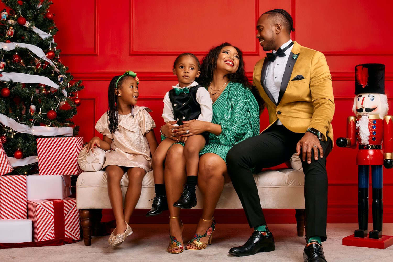 Family poses for holiday photos by decorated Christmas tree with red backdrop and wrapped presents.