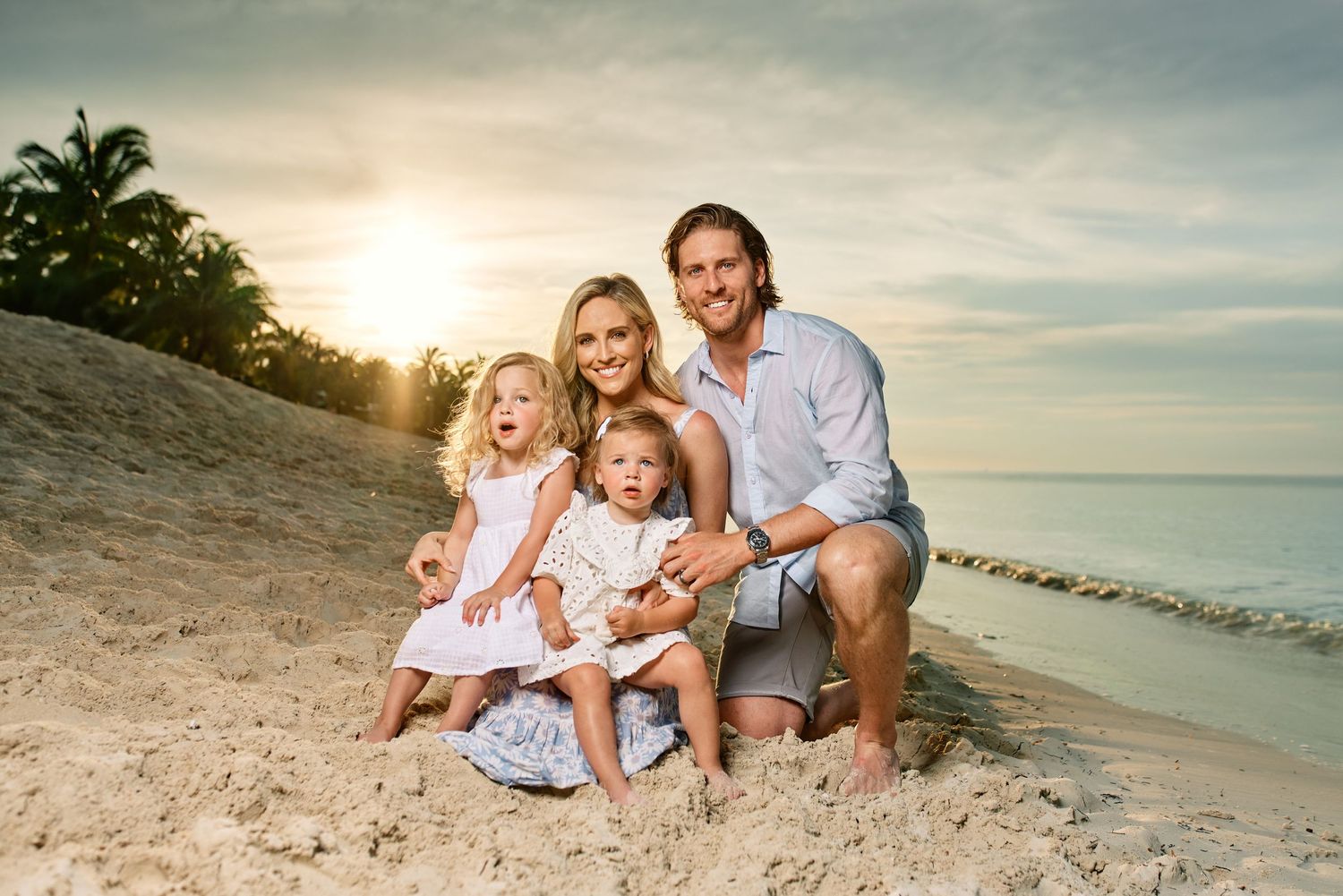Family sitting together on beach at sunset in coordinated light colored outfits.