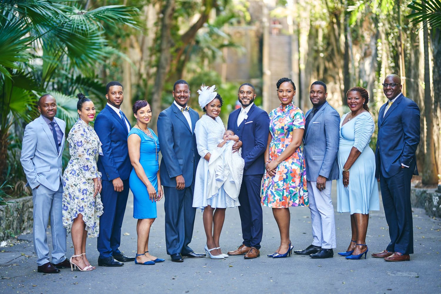 Formal group photo of wedding party members in blue and white attire on a palm-lined path.