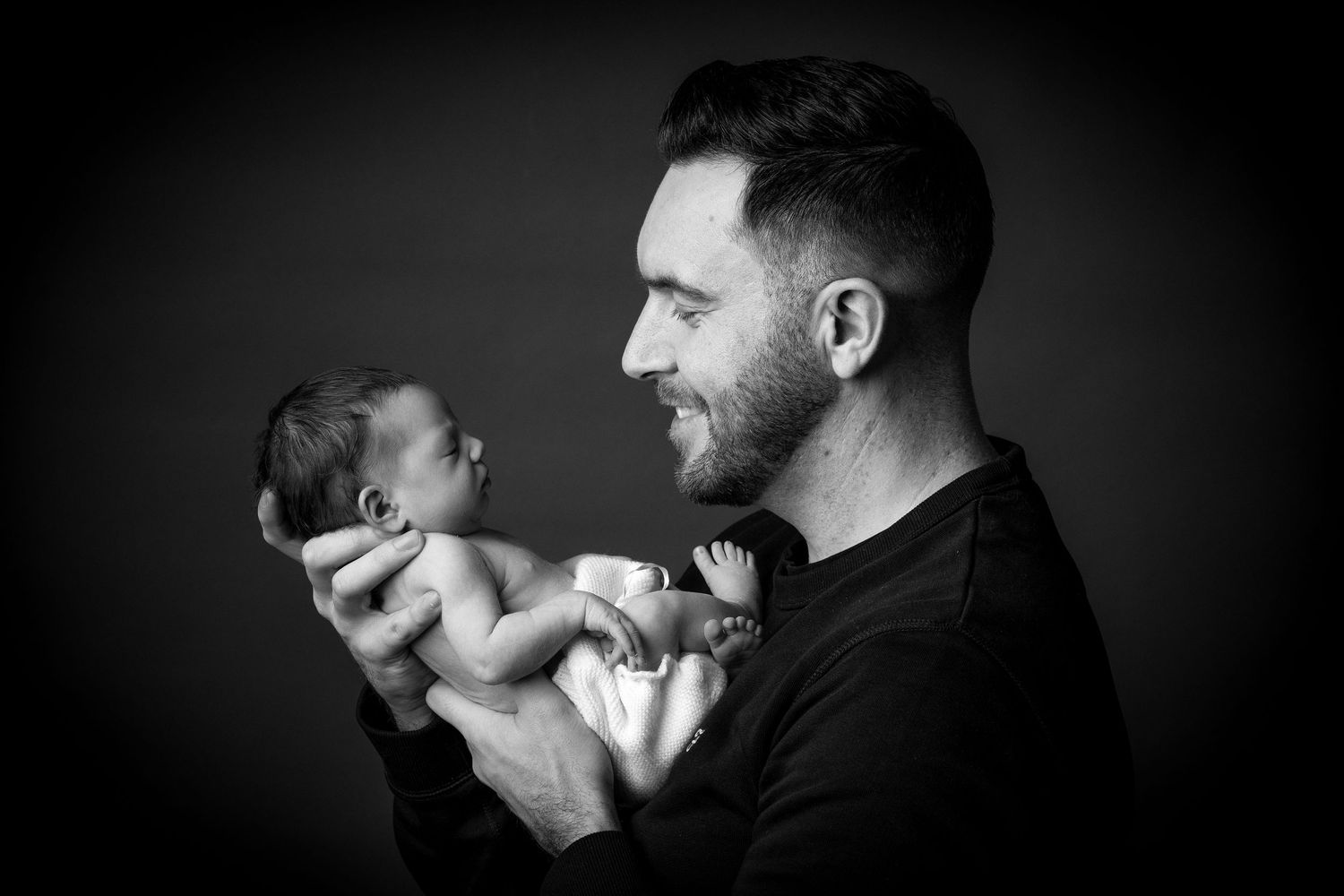 Black and white portrait of parent holding newborn baby against dark background.