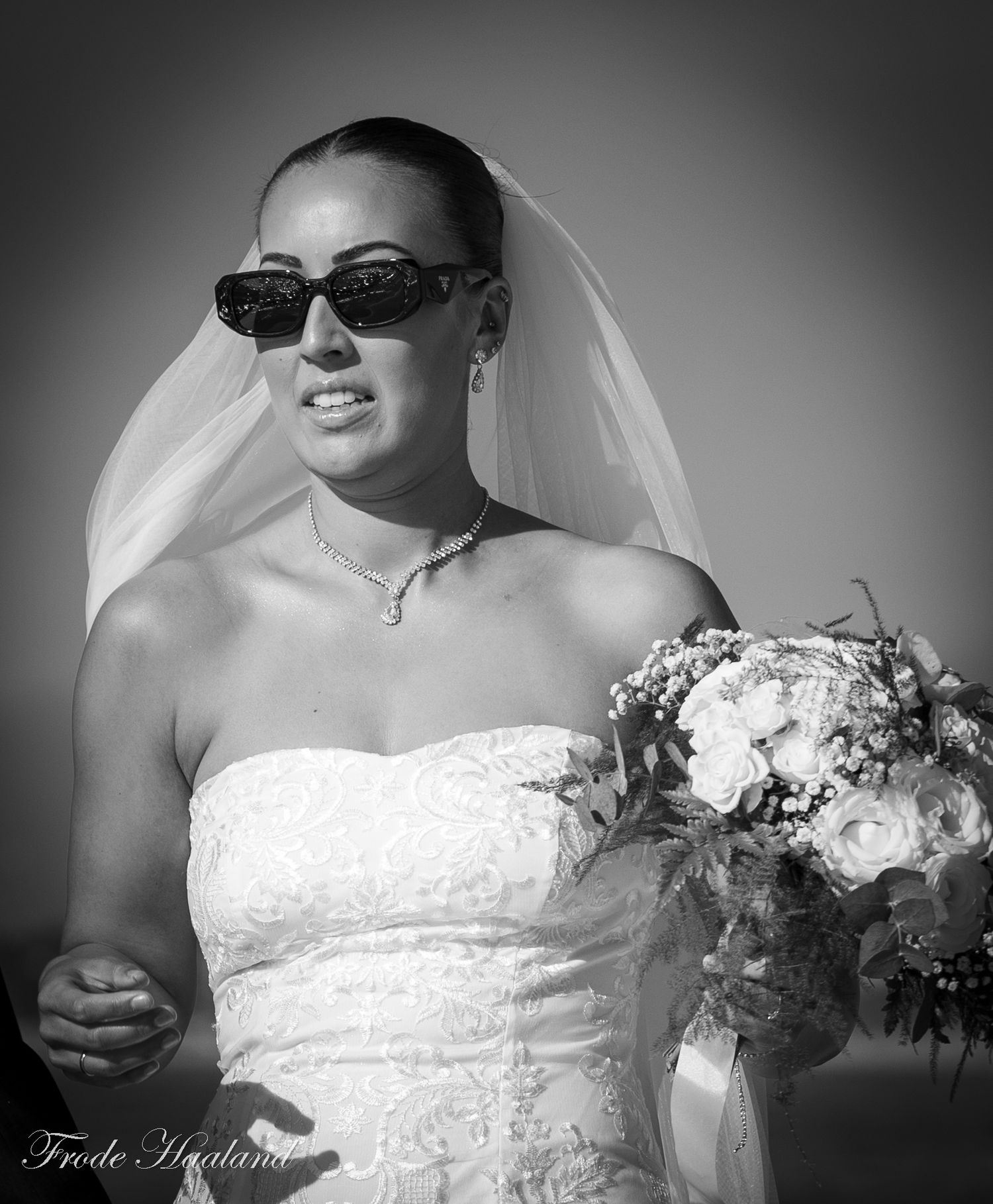 Black and white photo of a bride wearing sunglasses and a veil, holding a bouquet of roses.