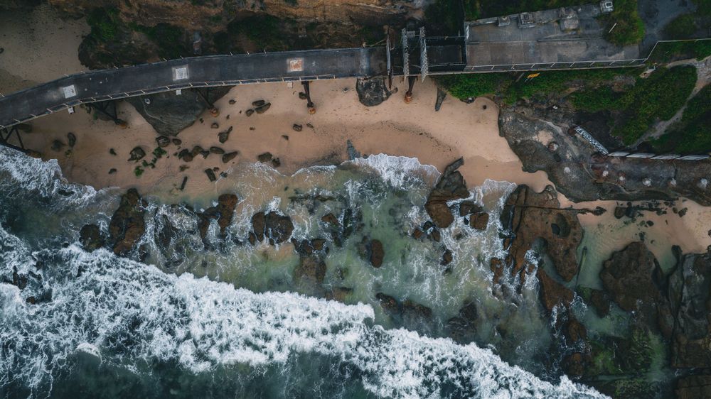 Bird's eye view of waves meeting rocky shore with sandy beach and coastal infrastructure.