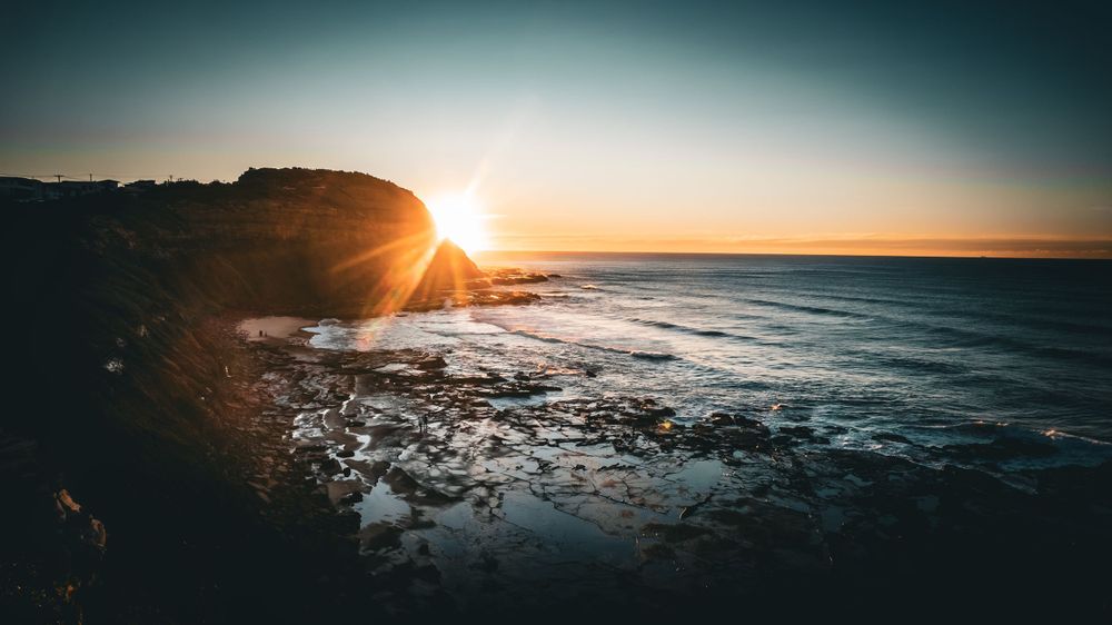 Golden sunburst peeks over coastal cliffs as waves crash against rocky shore at sunset.