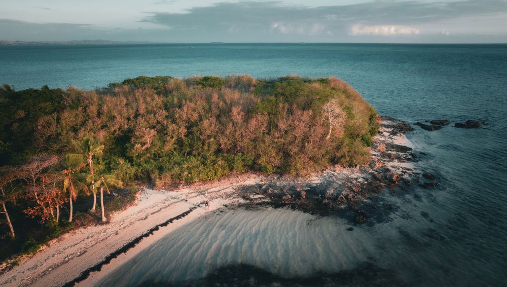 Drone shot of rocky peninsula with sandy beach and tropical vegetation against turquoise waters.
