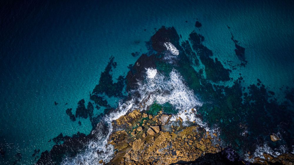 Aerial view of ocean waves crashing against rocky outcrop in turquoise waters.
