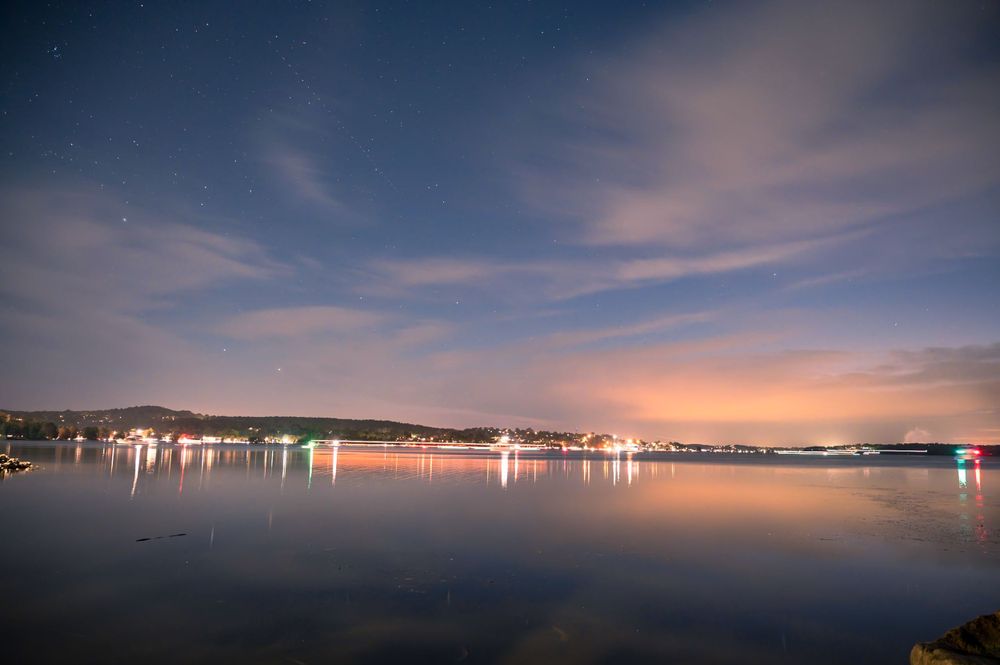 Night cityscape panorama reflected in calm waters with twinkling lights along the shoreline.