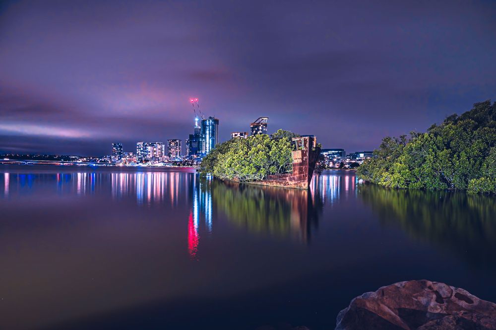 Purple twilight skyline reflected in still waters with city lights shimmering in the distance.