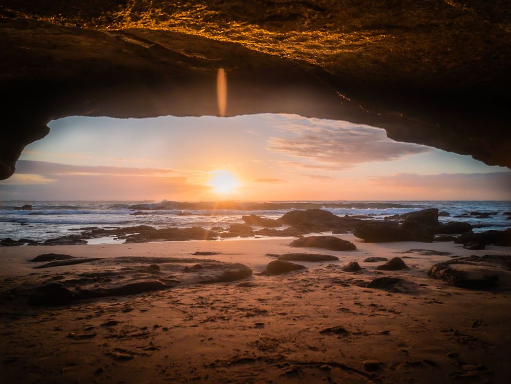 Dramatic ocean sunset viewed through a natural rock cave arch with waves crashing on rocky beach.