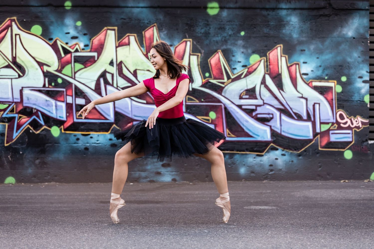 Ballet dancer in red and black leotard performs pose in front of colorful graffiti wall.