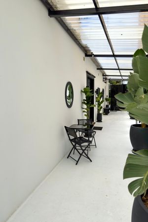 Modern minimalist corridor with white walls, translucent roof panels, black folding chairs and large potted plants.