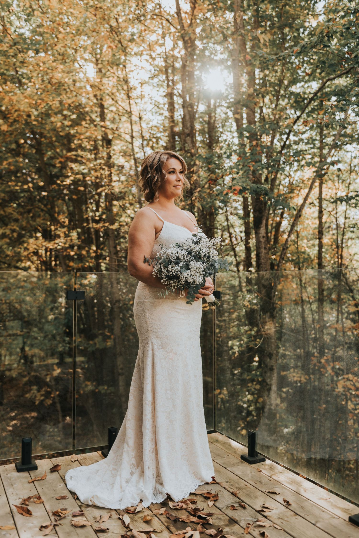 A bride in a fitted white dress holds a baby's breath bouquet while standing on a wooden deck surrounded by autumn trees.