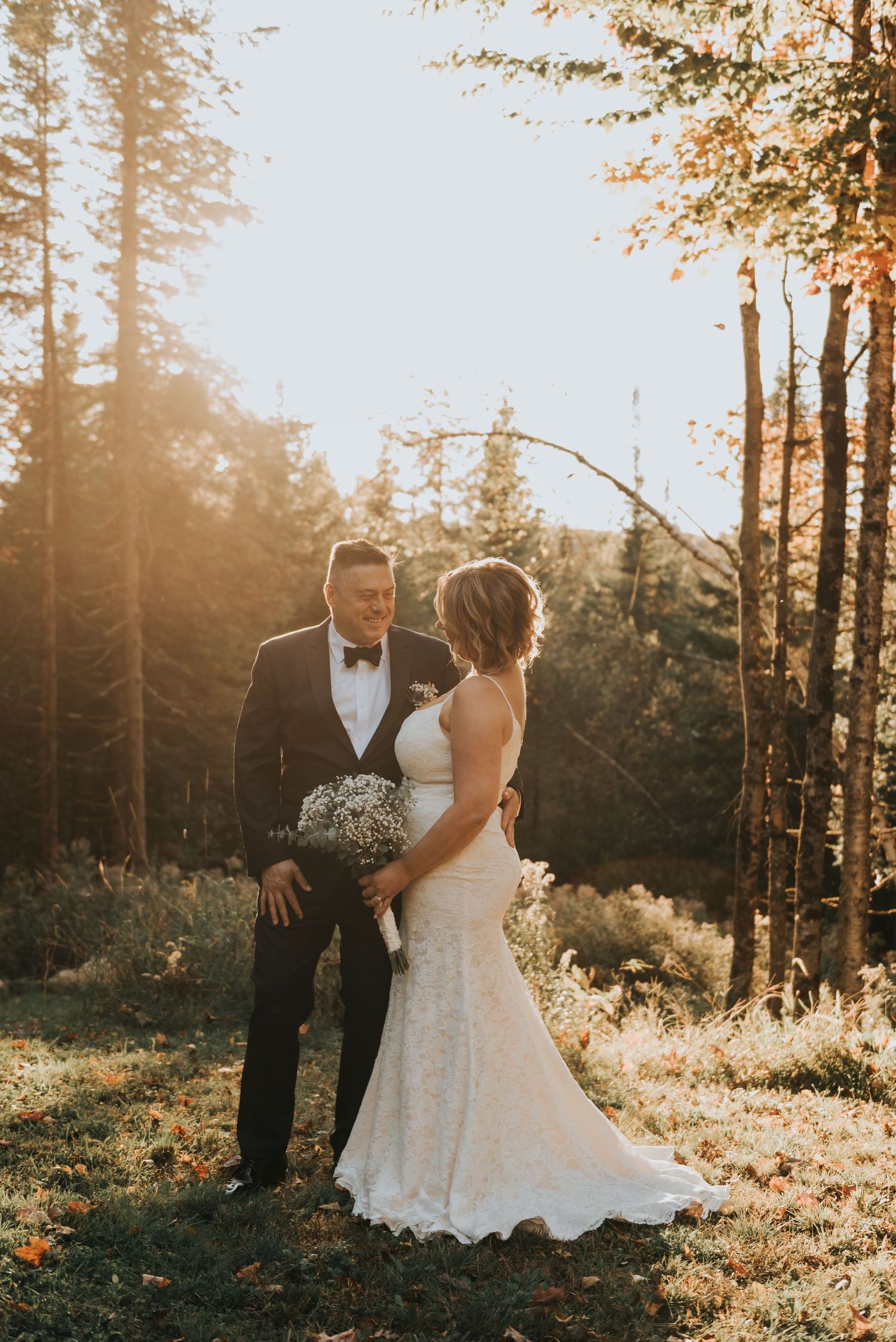 A romantic sunset wedding photo of a couple in formal attire standing among tall trees in a forest setting.