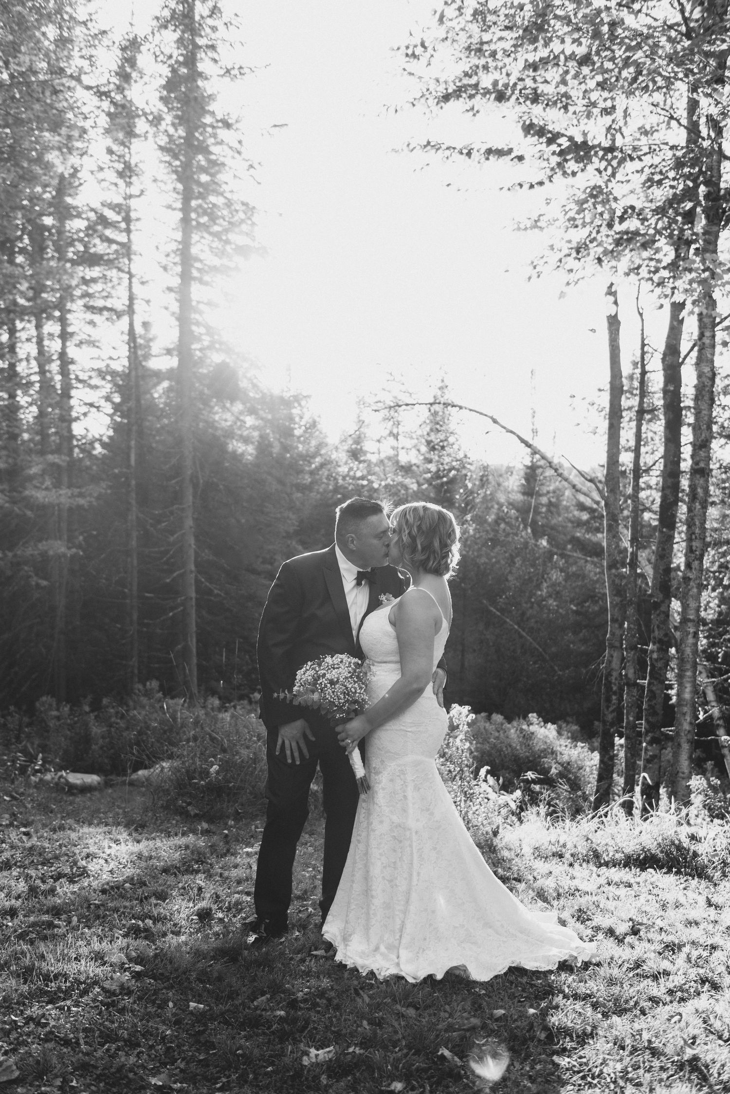 A romantic wedding couple shares a tender moment in a sunlit forest clearing, with sunbeams filtering through the trees.