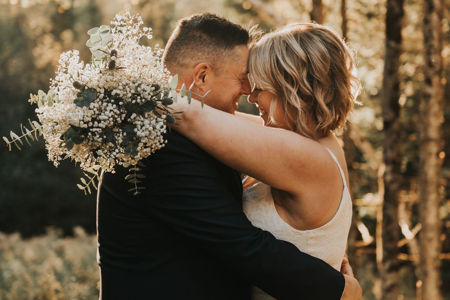 A romantic wedding couple embraces in a sunlit forest while holding a white floral bouquet.