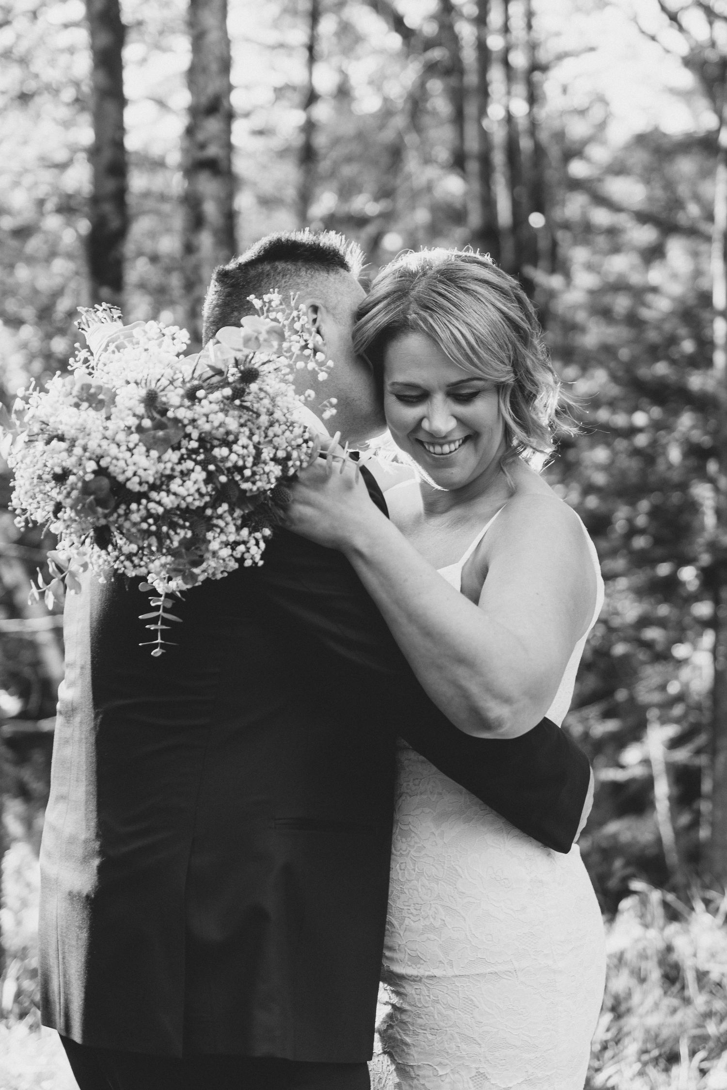 A couple share an intimate candid moment in the woods with a delicate bouquet of white flowers.