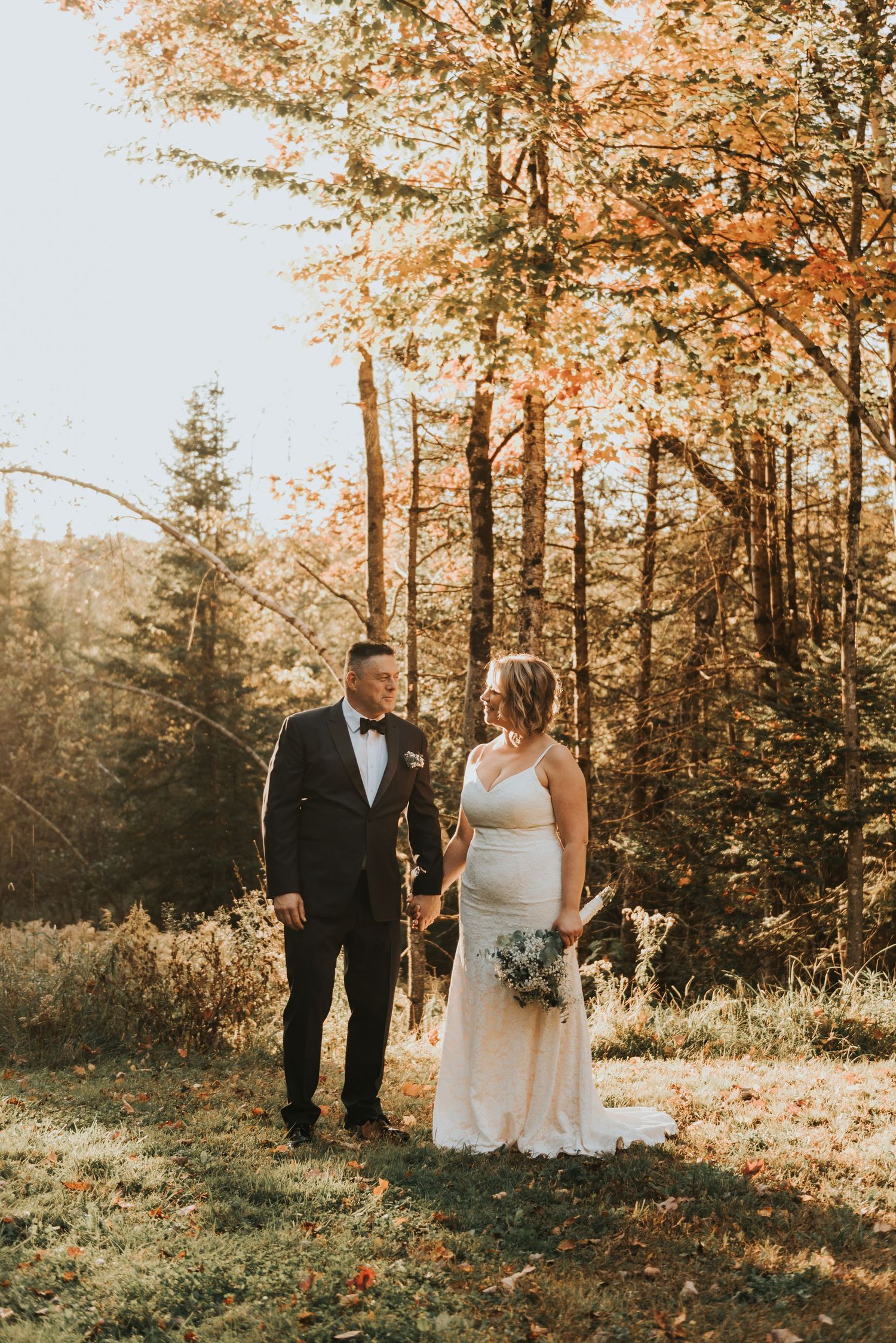 A couple stands together in an autumn forest setting with golden leaves and warm sunlight filtering through the trees.