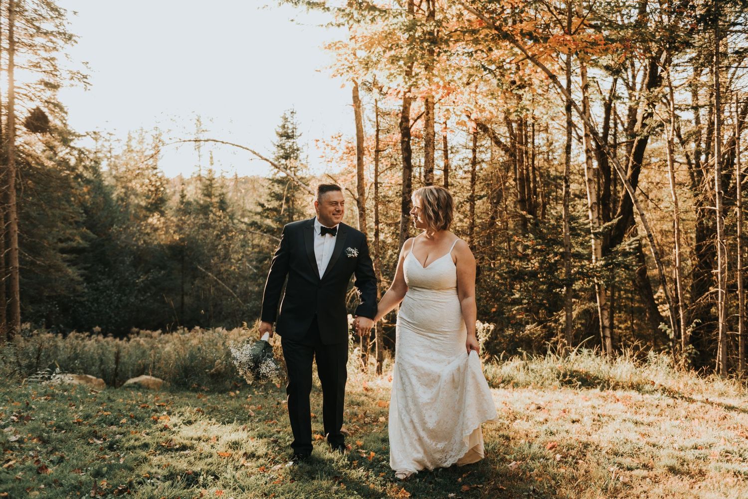 A couple holds hands while walking through a sunlit autumn forest during their wedding ceremony.