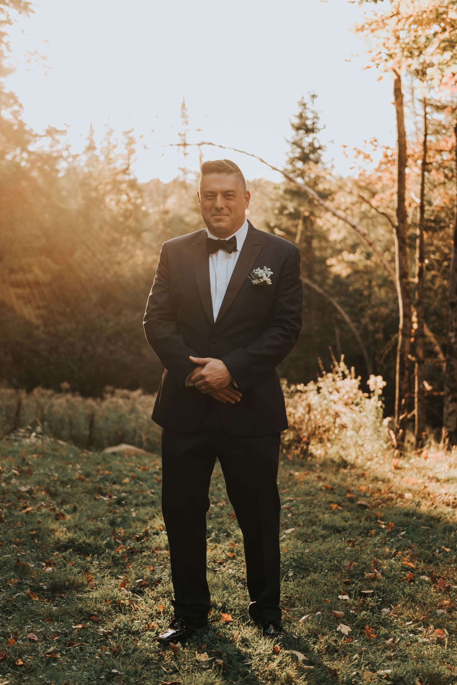 A groom in a black tuxedo and bowtie stands in golden sunlight against a wooded forest backdrop.