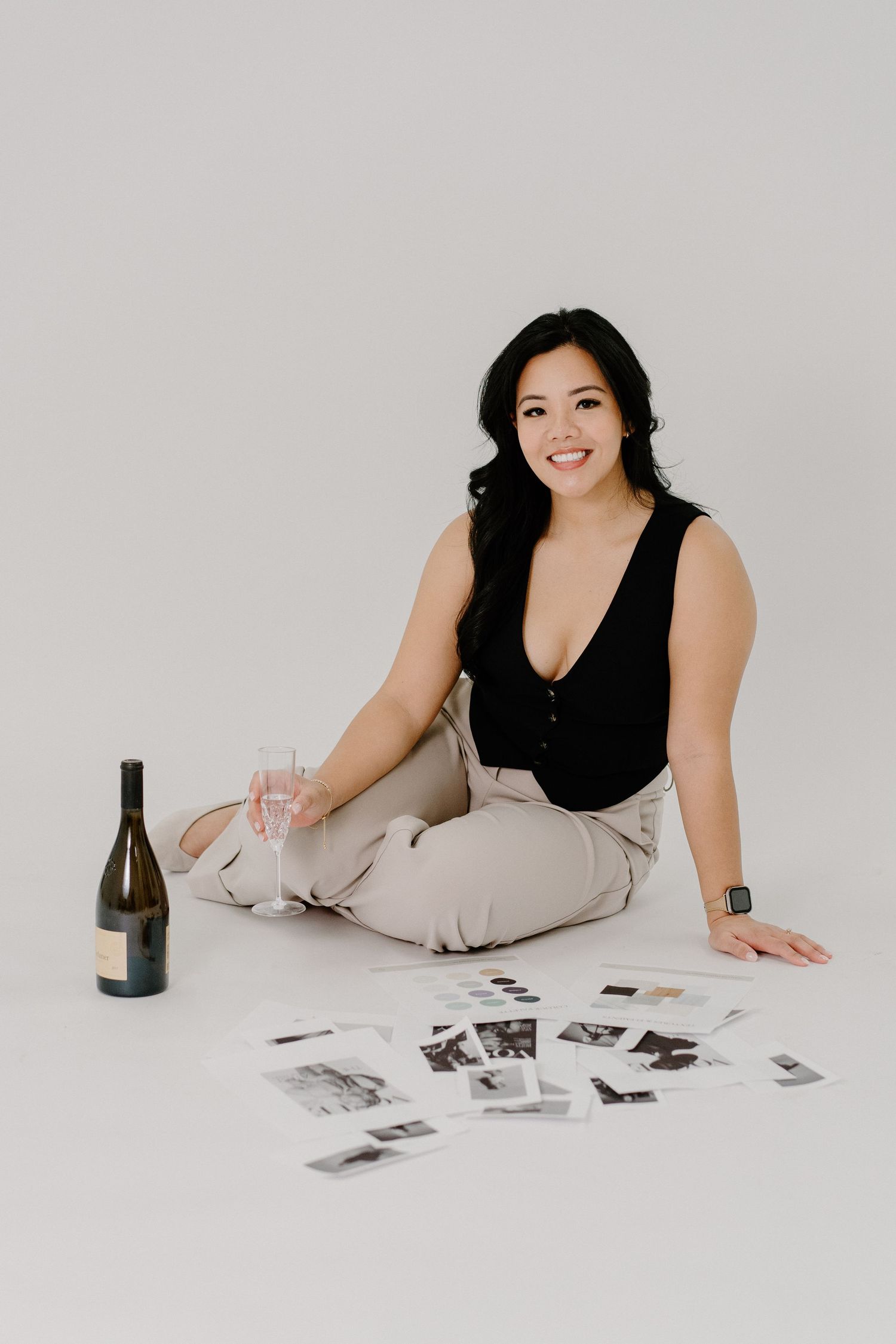 Lifestyle photo with black wine bottle and scattered photos on white floor while sitting in black top and beige pants.