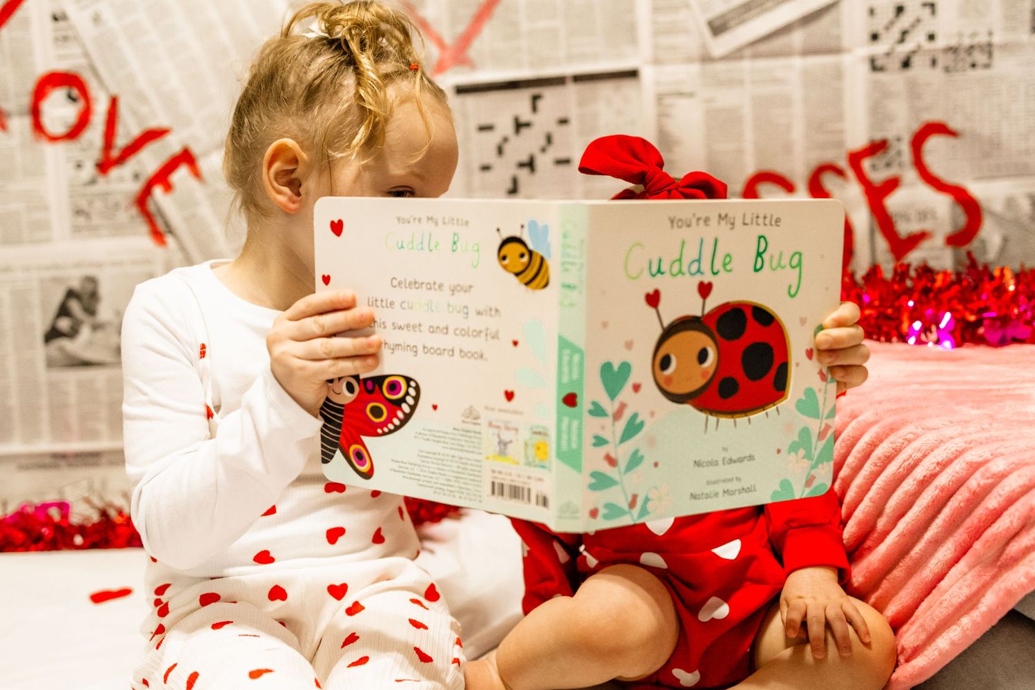 Small child in heart-printed pajamas reads a picture book against newspaper-covered wall with red love graffiti.