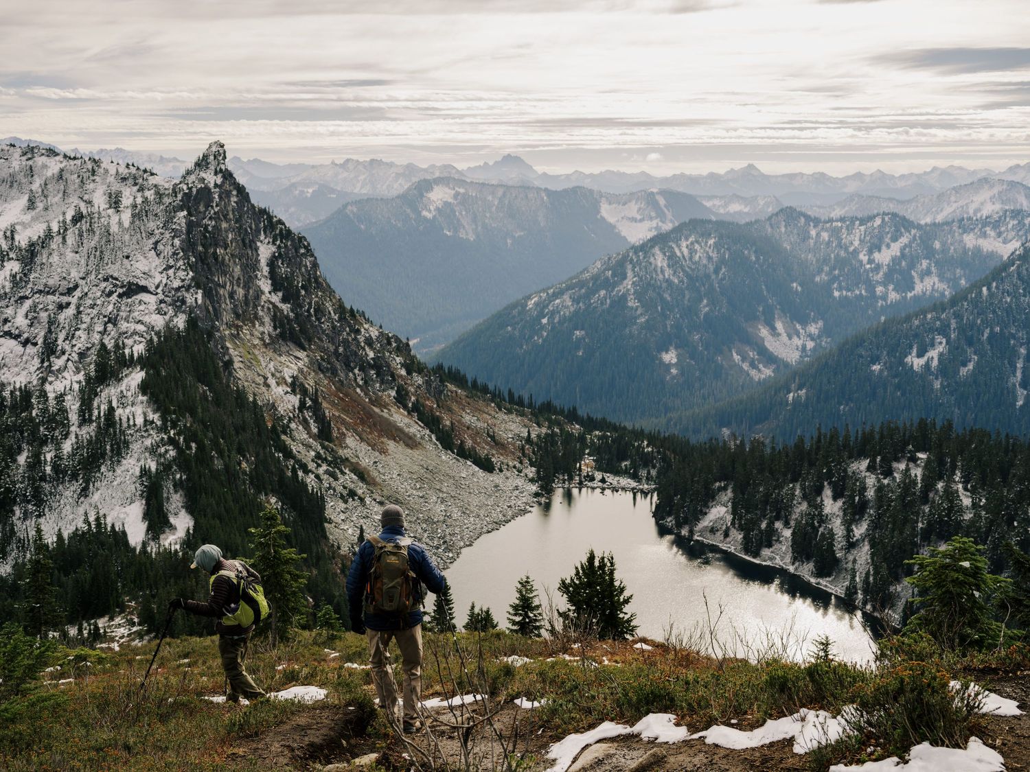 Two hikers stand on a mountain ridge overlooking a dramatic vista of snowy peaks in the Pacific Northwest.