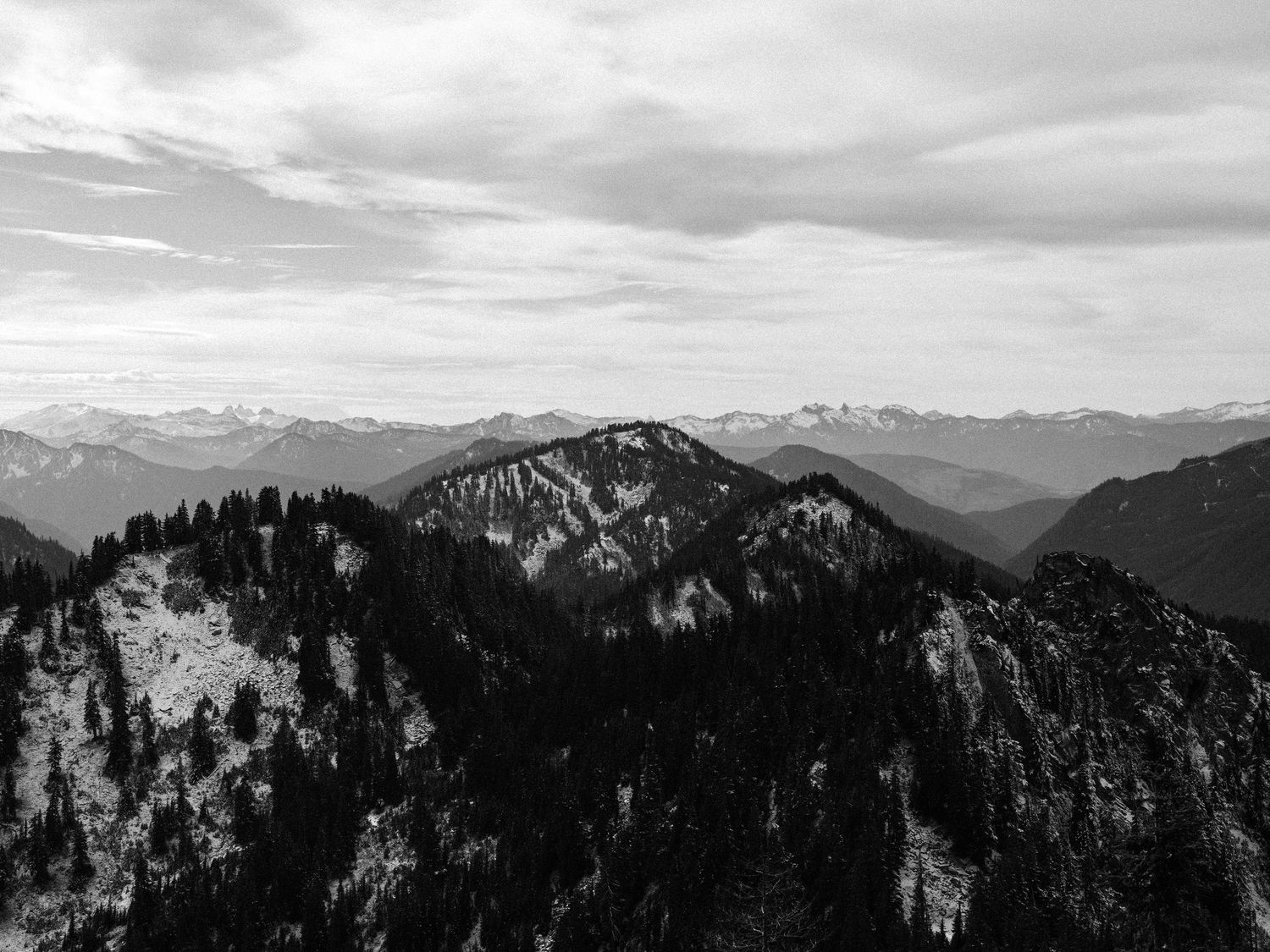 A dramatic black and white mountain landscape with snow-capped peaks and forested slopes under cloudy skies.