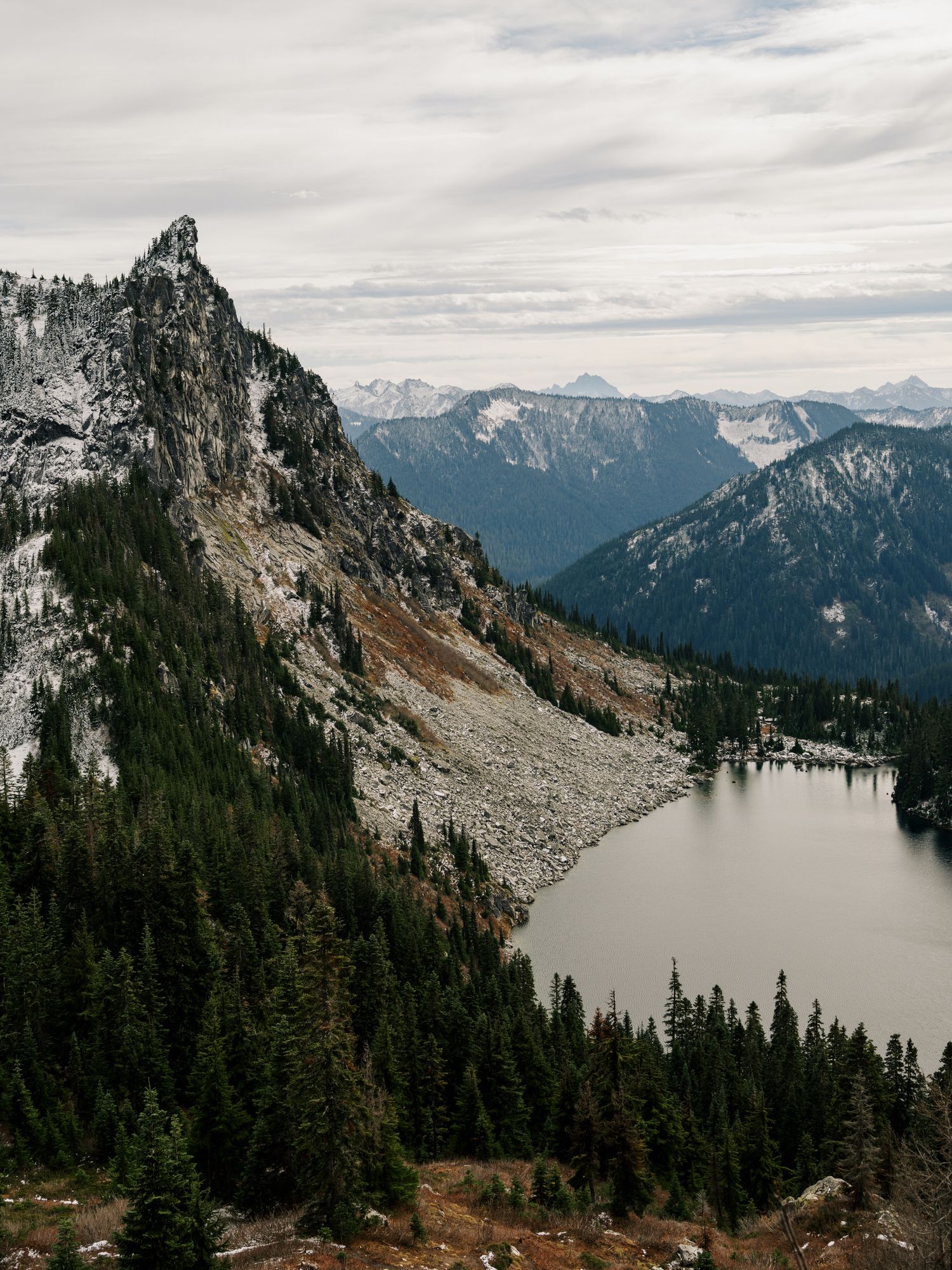 Dramatic mountain peak towers over an alpine lake surrounded by evergreen forest in misty Pacific Northwest wilderness.