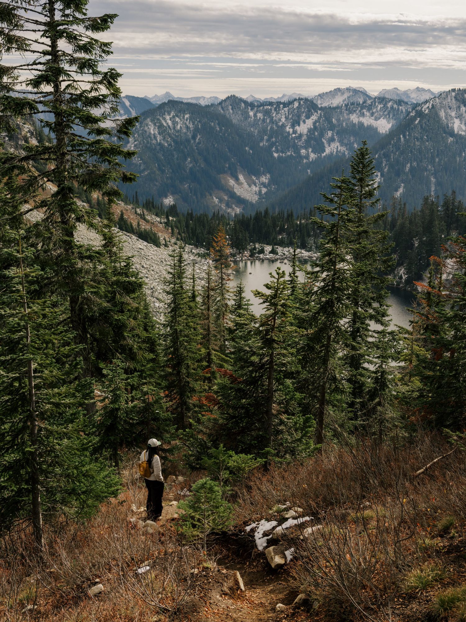 A hiker stands on a mountain trail overlooking evergreen forests and snow-capped peaks in the Pacific Northwest.