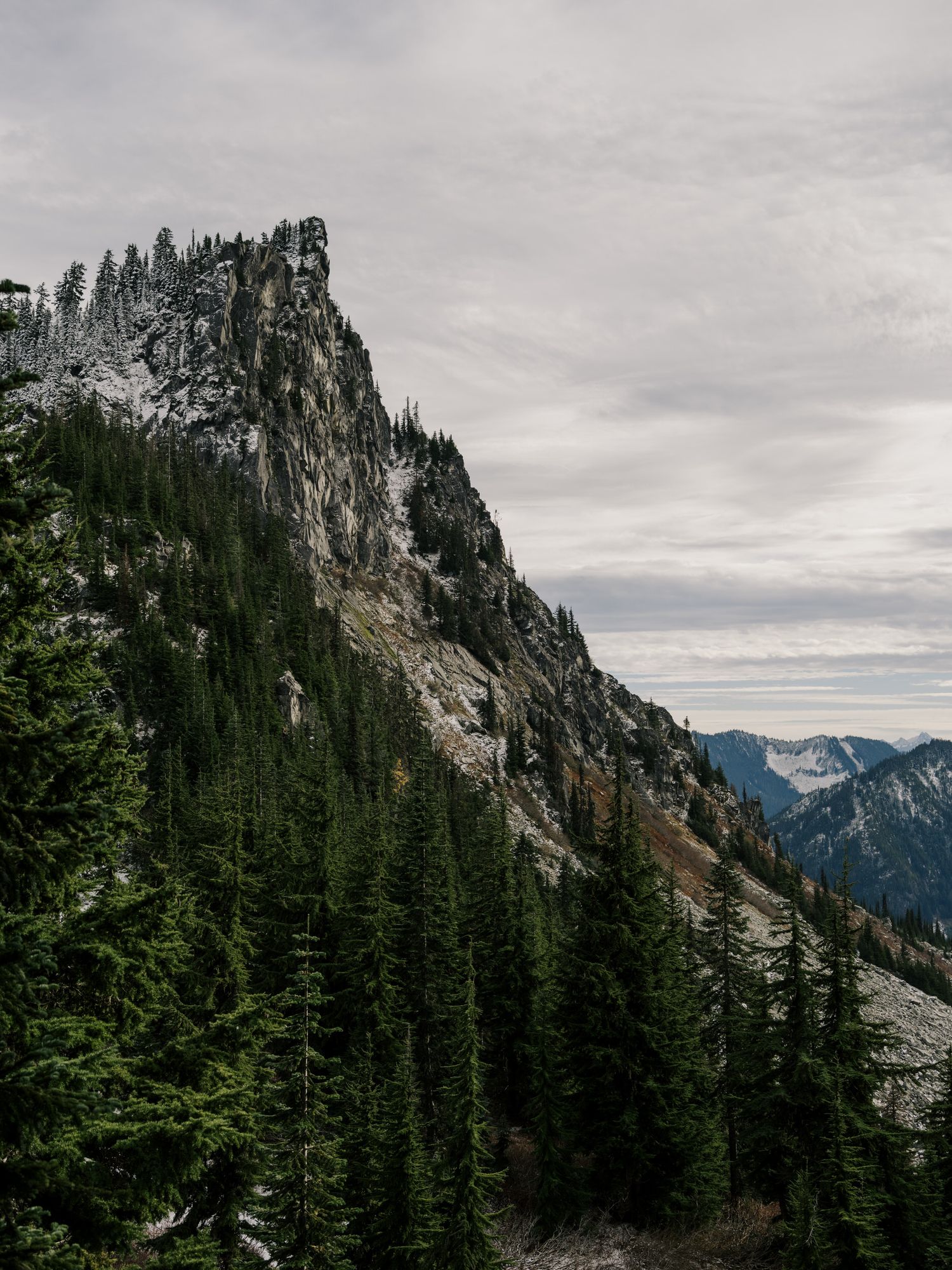 Dramatic rocky mountain peak towers over evergreen forest under moody gray skies in the Pacific Northwest wilderness.
