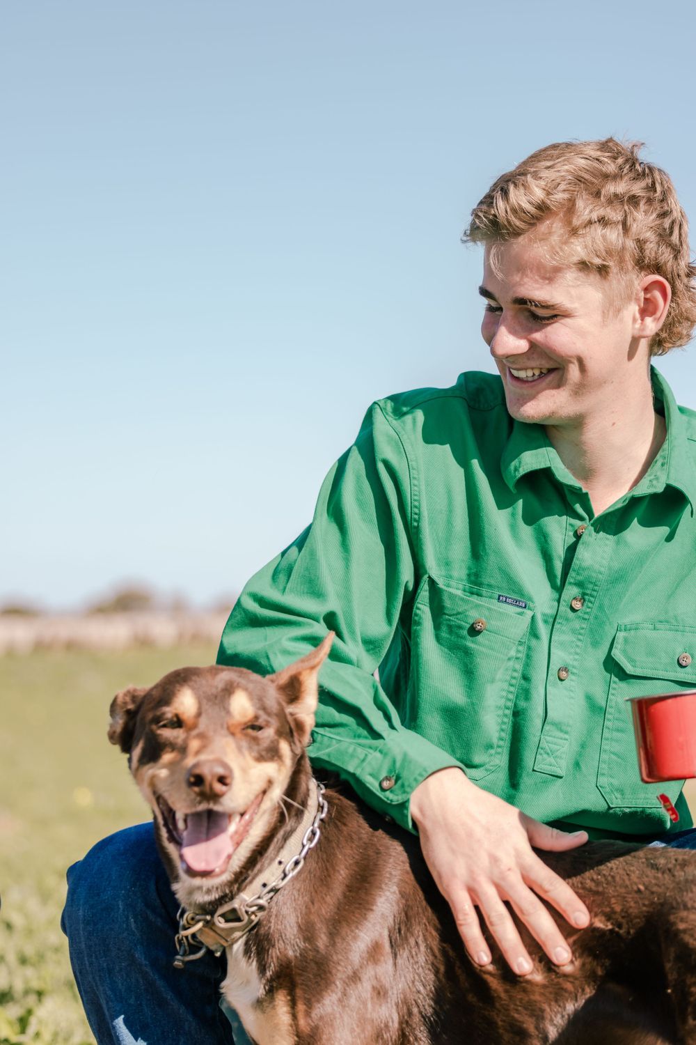 Person in green shirt smiles while petting happy brown dog outdoors.