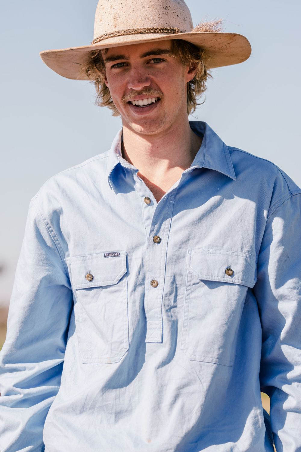 A person in a light blue work shirt and wide-brimmed hat stands outdoors against a bright sky.
