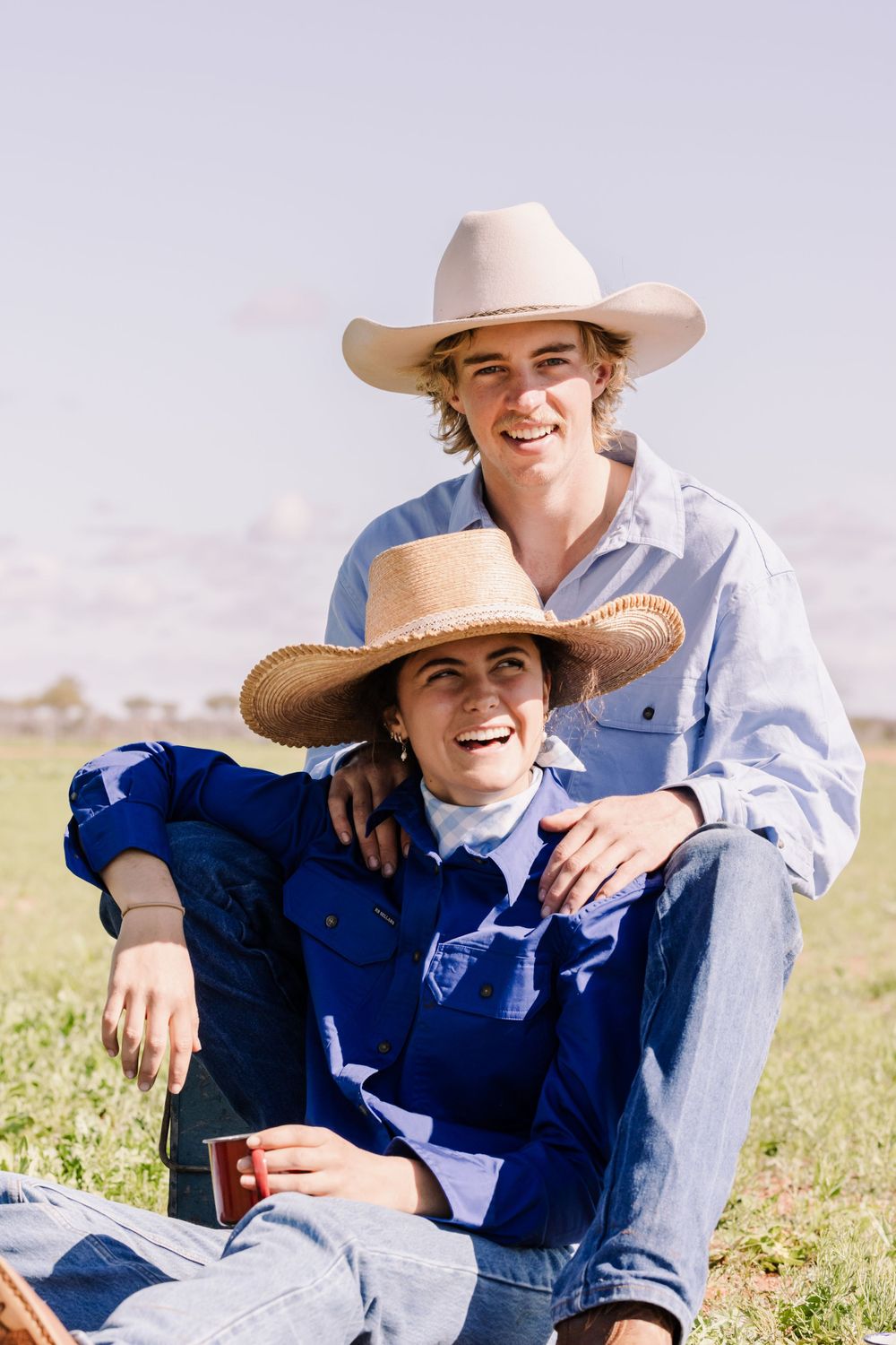 Two people wearing cowboy hats and denim relax together in a sunny field.
