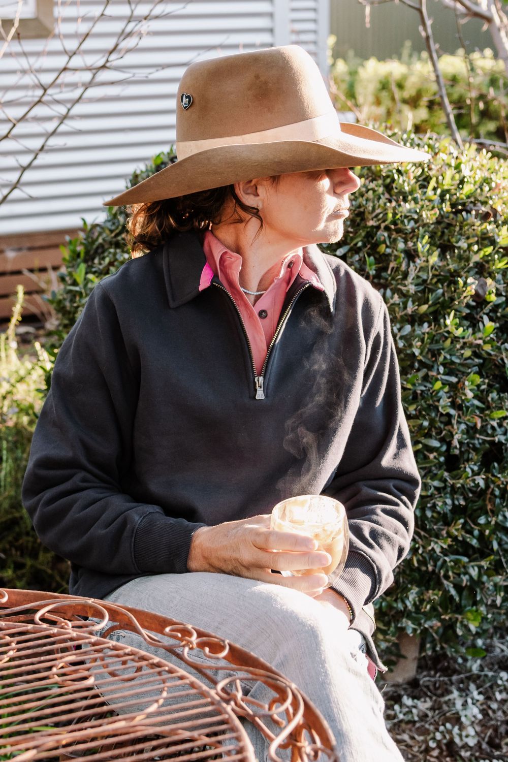 Person in cowboy hat and navy jacket enjoys coffee while relaxing outdoors.