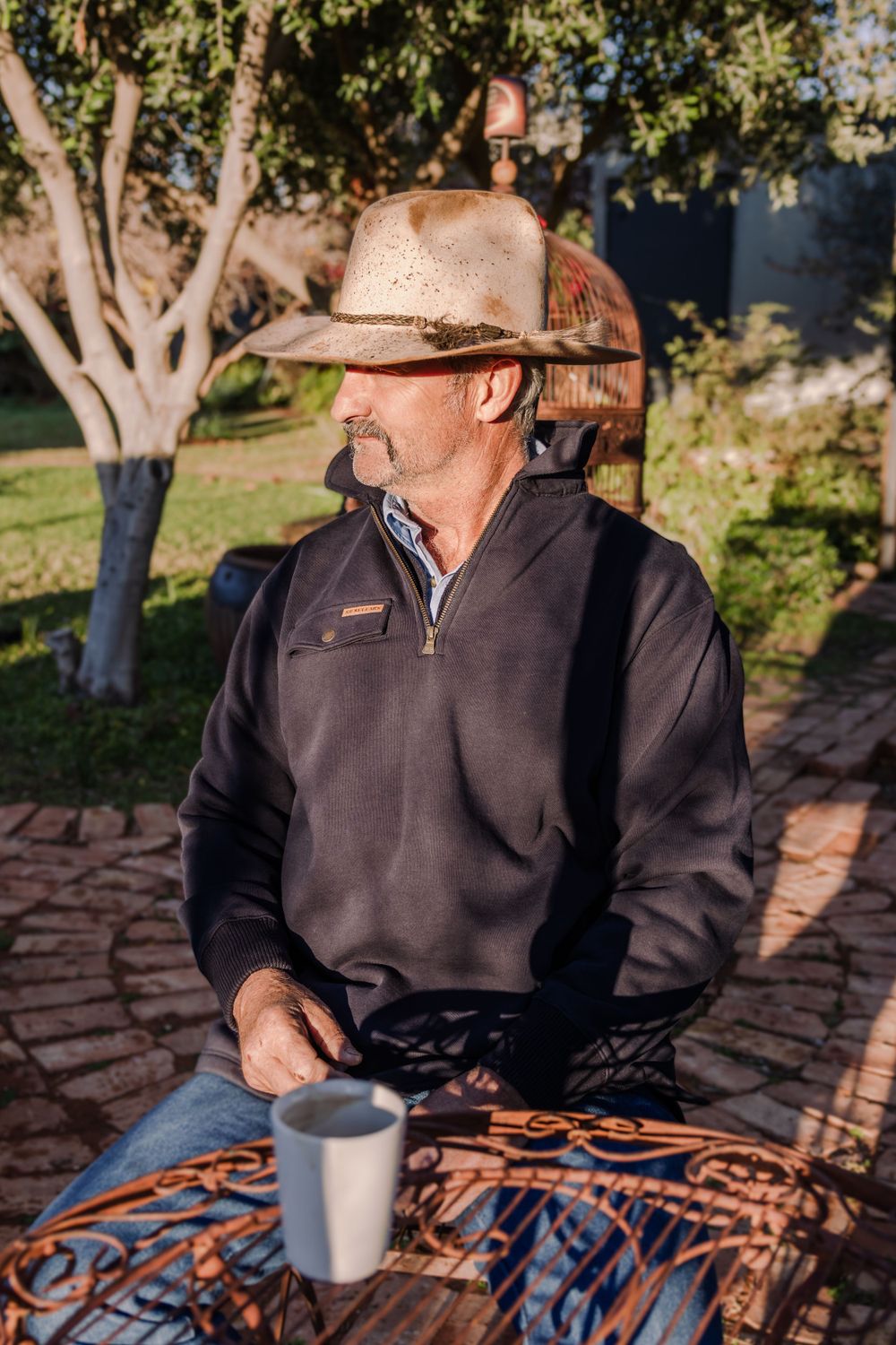 Person wearing cowboy hat and fleece pullover sits with coffee cup at outdoor table.